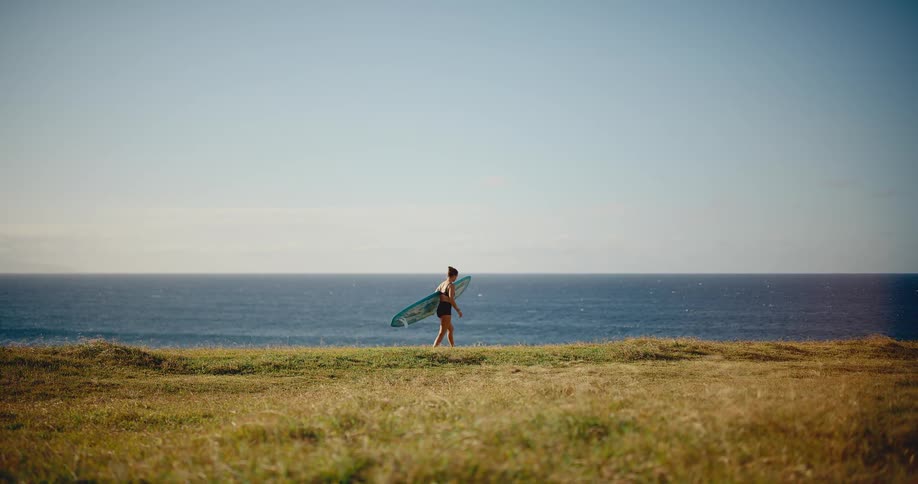 A female surfer walks along a grassy ridge overlooking the vast blue ocean on a beautiful sunny day