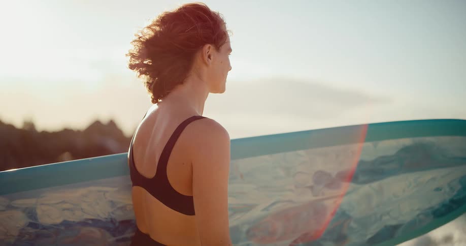 A serene surfer girl with her surfboard watches the ocean waves at golden hour in slow motion