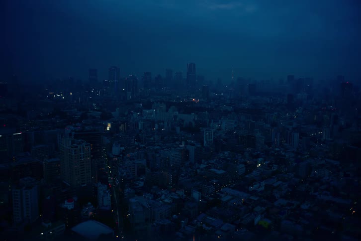 A vast aerial view of a dark and hazy cityscape at dusk, with twinkling city lights under a deep blue sky