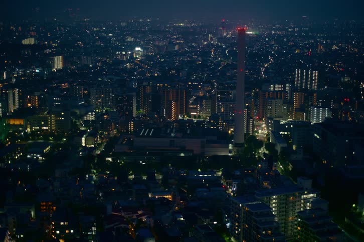 High-angle static shot of a vast cityscape at night, with countless lights illuminating the buildings and a tall industrial smokestack at the center