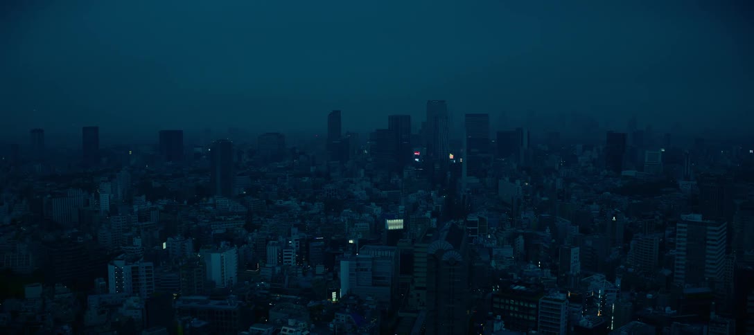 A sprawling city skyline is shrouded in a dark, blue haze at night, with only a few lights flickering in the distance