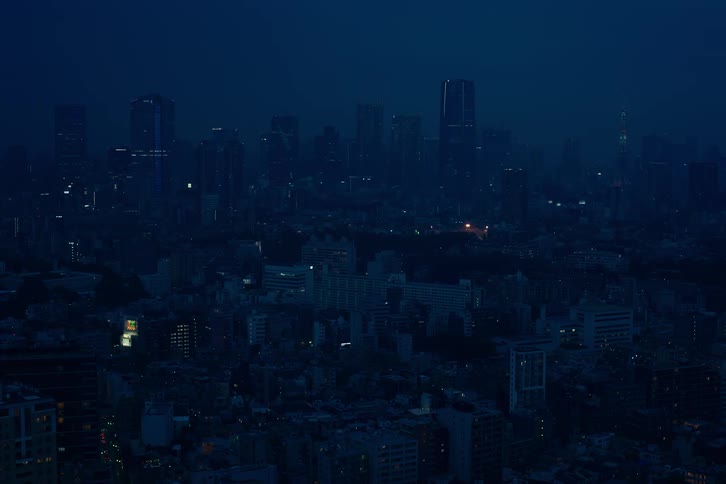 A wide, static view of a vast cityscape at night, cloaked in deep blue darkness with distant city lights twinkling