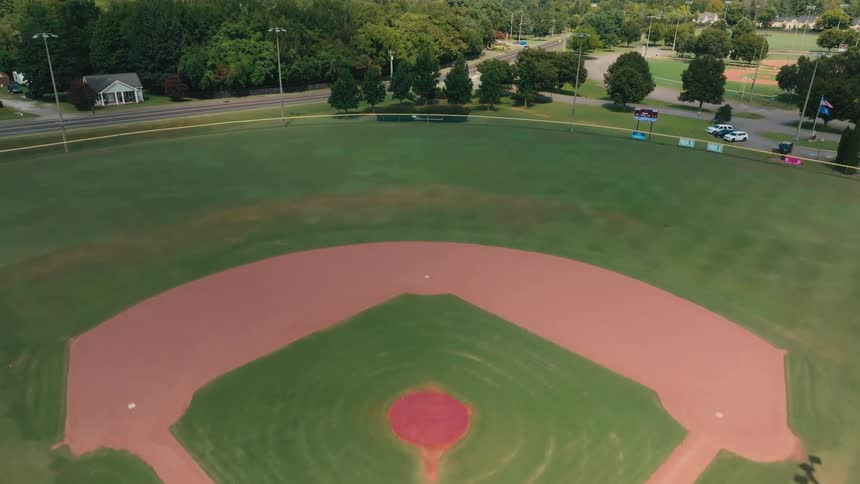 A smooth drone shot rises up and over the backstop of an empty baseball field in a community park