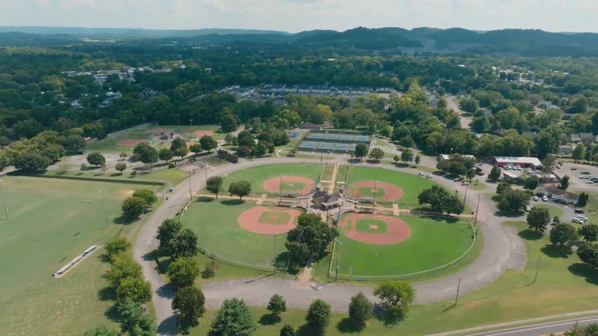 A slow aerial pullback reveals a large, green community park with four perfectly arranged baseball fields, tennis courts, and a soccer field on a sunny day