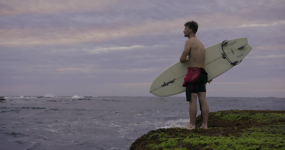 A man holds his surfboard looking at the water