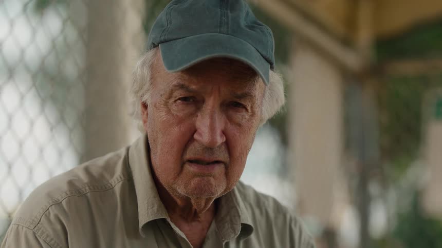 A pensive elderly man wearing a cap slowly looks up with a worried expression on his face