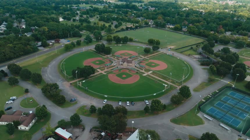 A slow aerial drone shot pushes in on a symmetrical, empty five-field baseball complex within a large community park