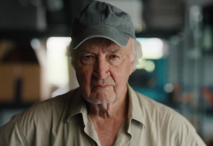 A close-up portrait of an elderly man with white hair and a stern expression, staring intensely into the camera from his workshop