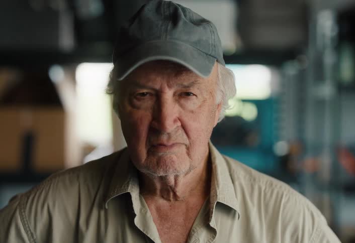 Portrait of a serious elderly man wearing a cap, looking directly at the camera in a dimly lit workshop