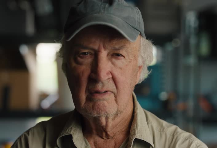 A serious elderly man in a hat slowly looks up into the camera with an intense gaze while in a workshop