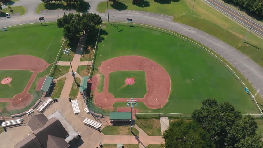 A slow aerial ascent from a top-down view reveals an empty baseball park complex on a bright, sunny day