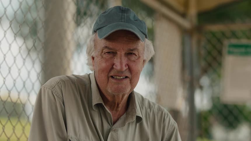 Close-up portrait of a smiling senior man wearing a cap, sitting by a chain-link fence at a park