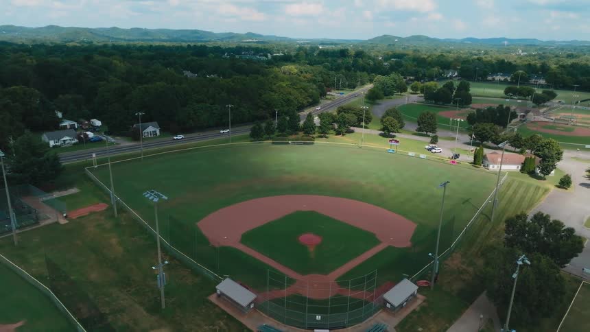 An aerial drone shot slowly pulls back to reveal an empty baseball field at a park on a sunny day