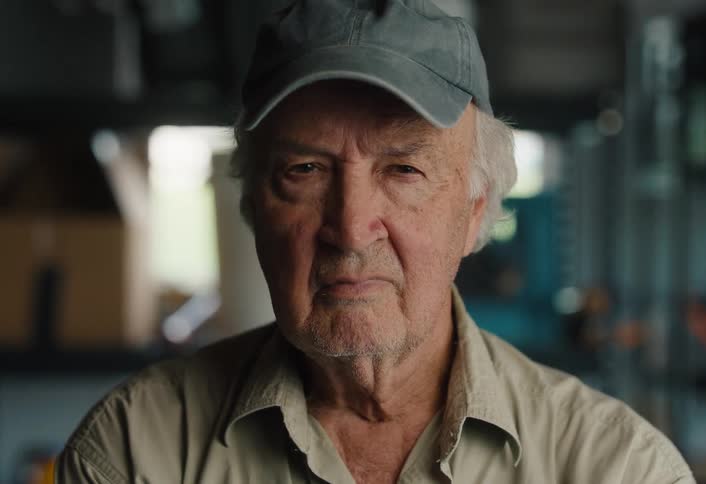 An elderly man in a cap slowly looks up with a serious and intense expression in his workshop
