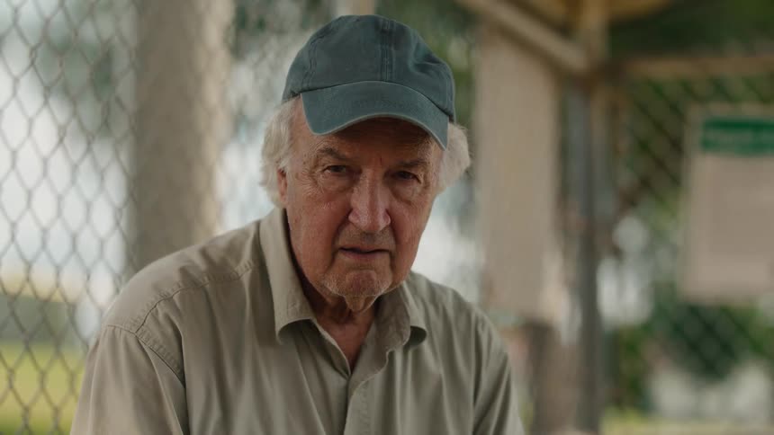 An emotional elderly man in a baseball cap looks up from his thoughts with a sad expression into the camera