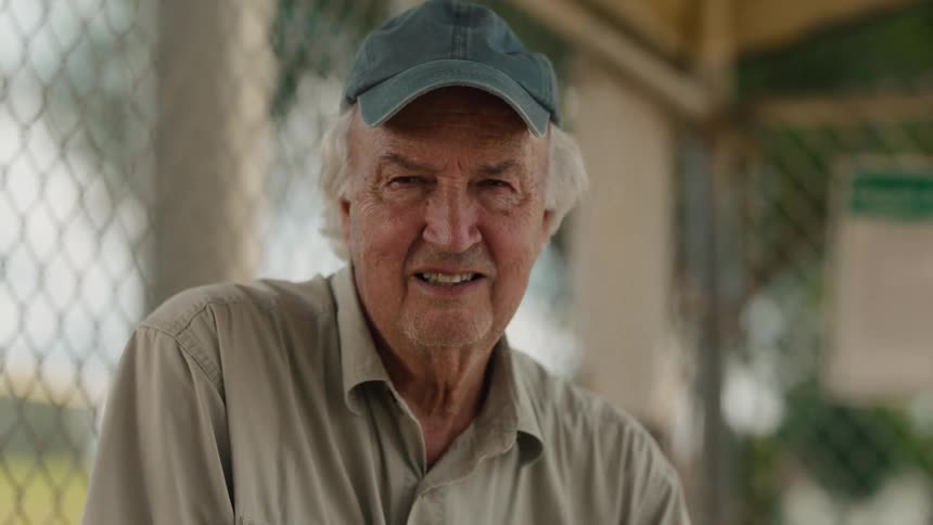 An elderly man in a cap sitting by a fence looks over and gives a kind smile to the camera in slow motion
