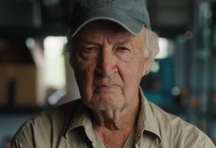 An elderly man in a baseball cap slowly raises his head and stares intently into the camera in his workshop