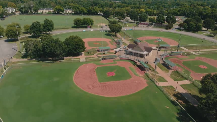 A slow aerial approach towards empty baseball diamonds in a suburban park on a bright, sunny afternoon