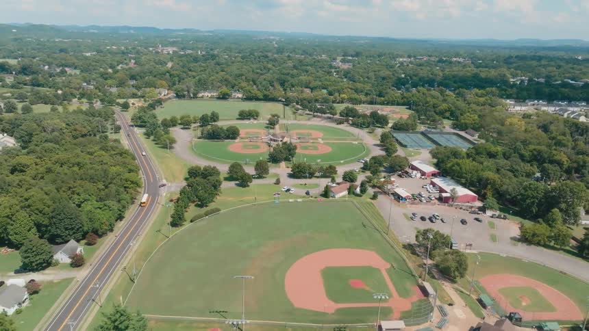 An aerial pullback shot reveals a large community sports complex featuring multiple baseball diamonds, a football field, and tennis courts on a bright, sunny day