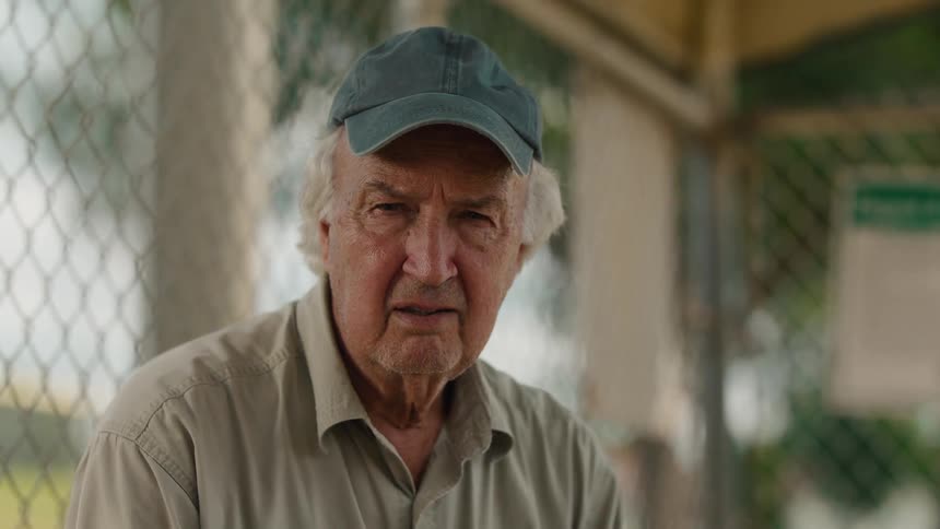 A portrait of an elderly man with a thoughtful expression sitting in a dugout, who then looks at the camera and offers a gentle smile