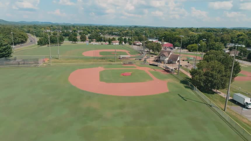 A slow aerial fly-in over an empty community sports complex featuring multiple baseball diamonds and a football field on a sunny day