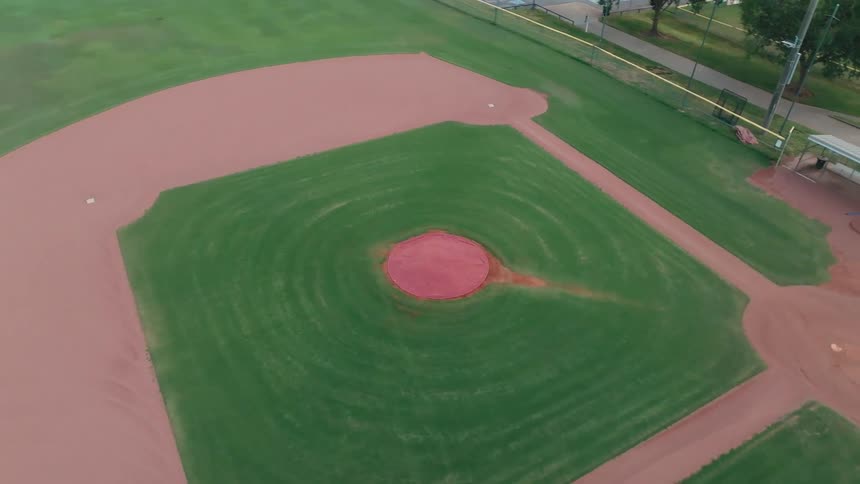 An aerial view rises high above an empty, well-manicured baseball field, revealing the diamond, dugouts, and surrounding park