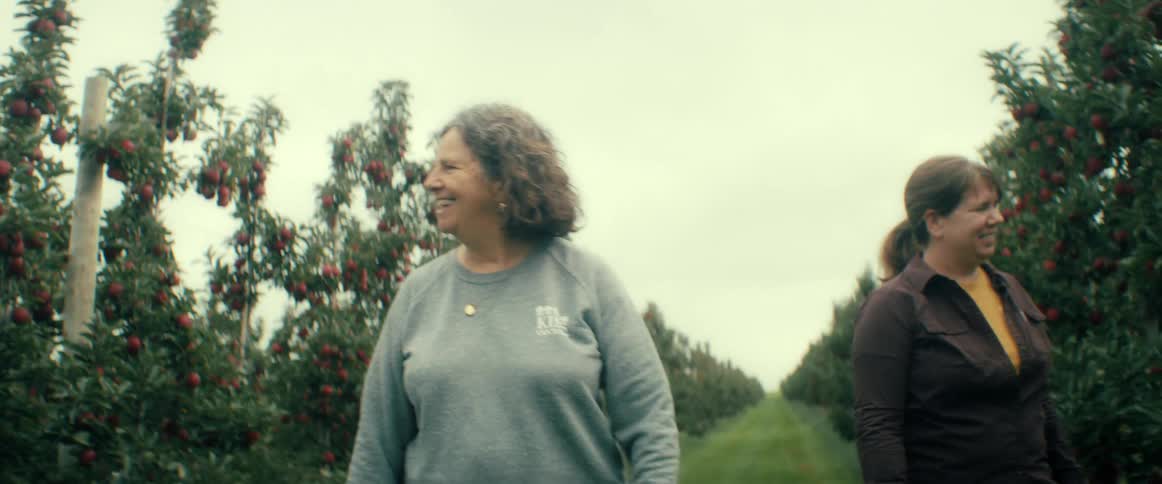 Two smiling women happily walk through an apple orchard filled with ripe red apples