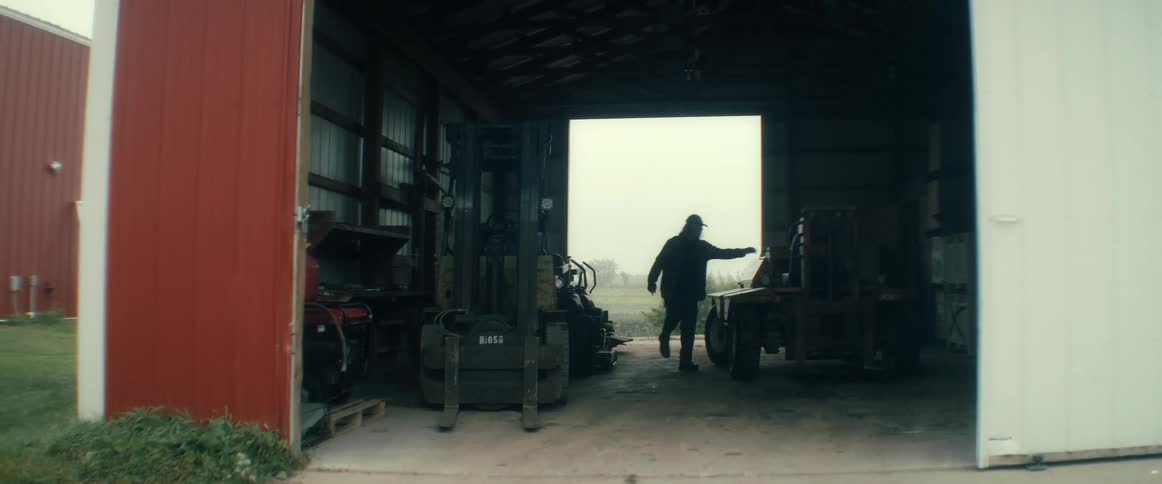 A silhouetted worker walks through a dark farm garage and climbs onto a piece of heavy machinery on an overcast day