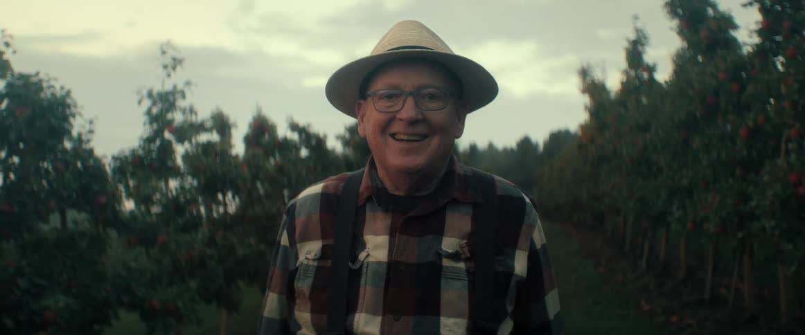 A friendly senior farmer in a straw hat smiles proudly at the camera in his apple orchard