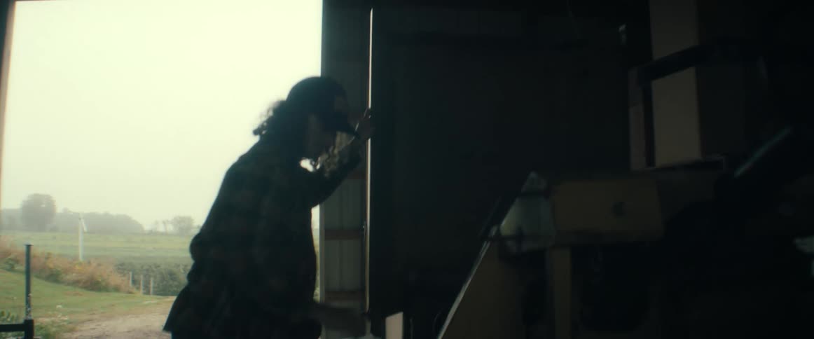 A person in a flannel shirt and hat closes a large barn door, picks up a bundle of twigs, and tosses them out into the misty rural landscape