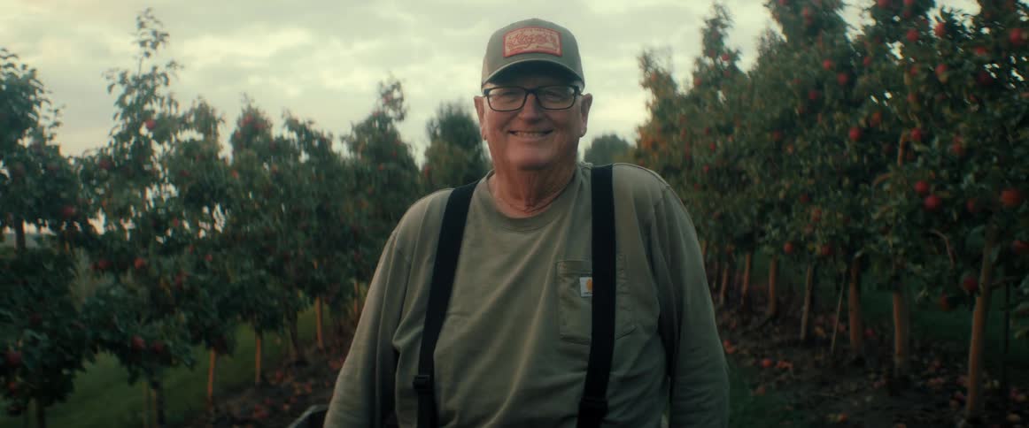 A friendly, elderly farmer smiles warmly for the camera while standing in his apple orchard