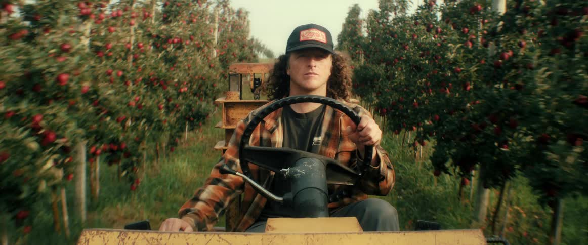 A young farmer with long curly hair drives a tractor through the rows of a vibrant apple orchard during harvest time