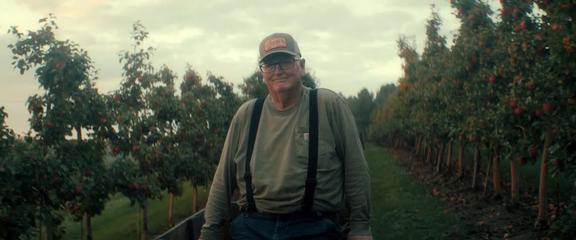 A proud elderly farmer with a warm smile sits in his apple orchard at dusk