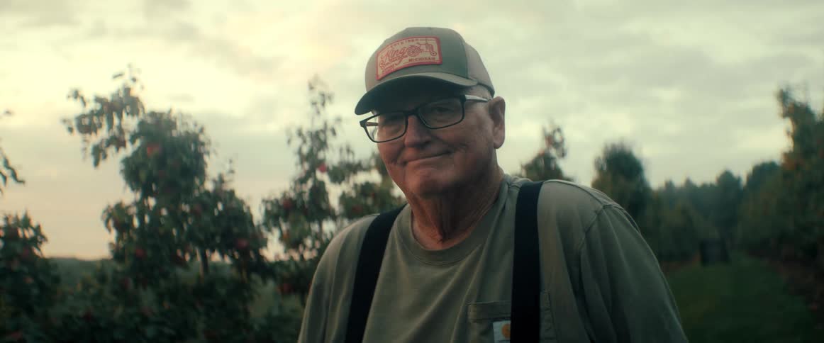 A proud, senior farmer wearing a cap and glasses turns to smile at the camera while standing in his apple orchard at dusk