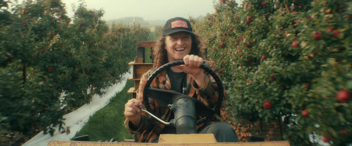 A happy young farmer with long hair drives a tractor through a beautiful apple orchard while looking and smiling at the camera