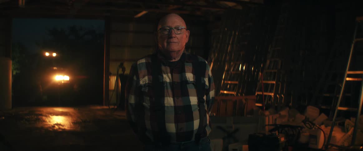 Portrait of a senior man in a plaid shirt standing inside a dark barn, looking up at the camera with a gentle expression