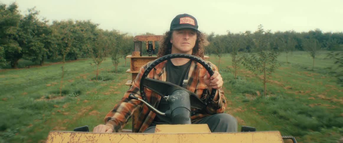 A man with long curly hair wearing a flannel shirt and hat drives a yellow farm vehicle through an orchard