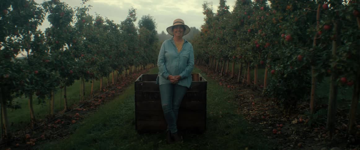 A smiling female farmer wearing a hat proudly stands between rows of apple trees in an orchard on a misty morning