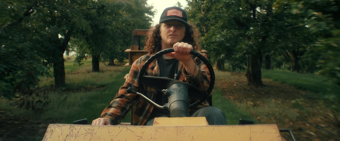 A young farmer with long curly hair wearing a plaid shirt drives a tractor down a grassy path through a lush green orchard
