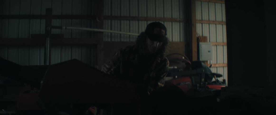 A focused woodworker with long hair uses a power router to carve a piece of wood in a dimly lit barn