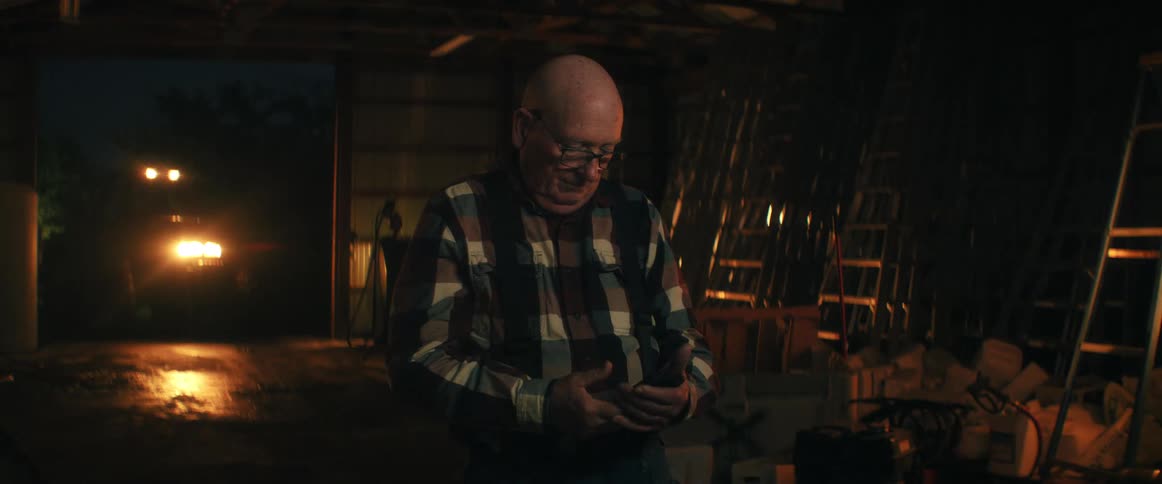A senior farmer in a plaid shirt happily ends a phone call and texts on his smartphone at night in his barn
