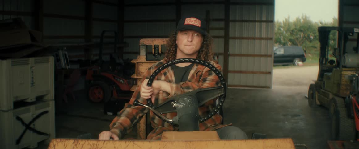 A young man with curly hair wearing a plaid shirt and baseball cap drives a forklift out of a large farm garage