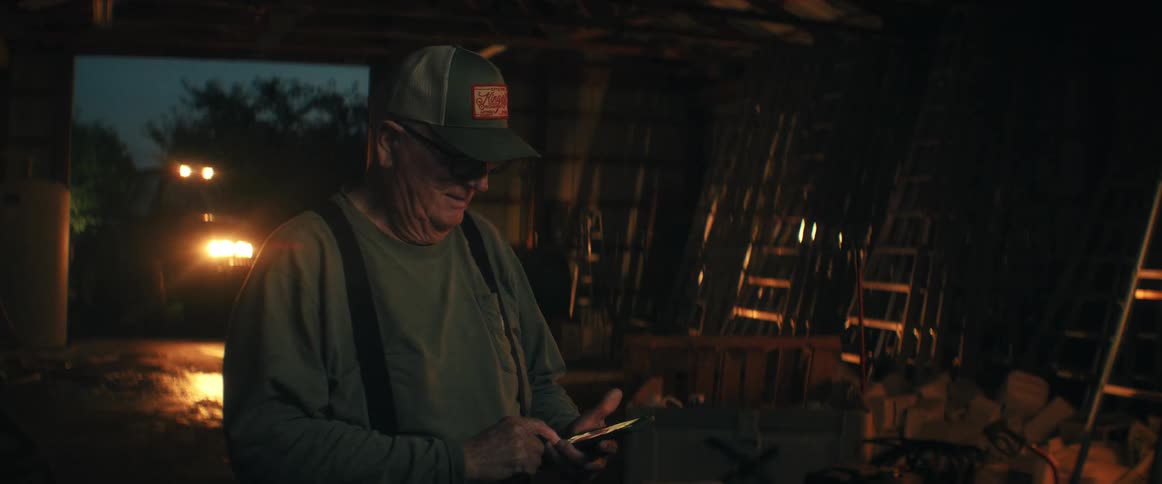 An older man wearing a cap and suspenders smiles while scrolling on his smartphone in a dimly lit workshop