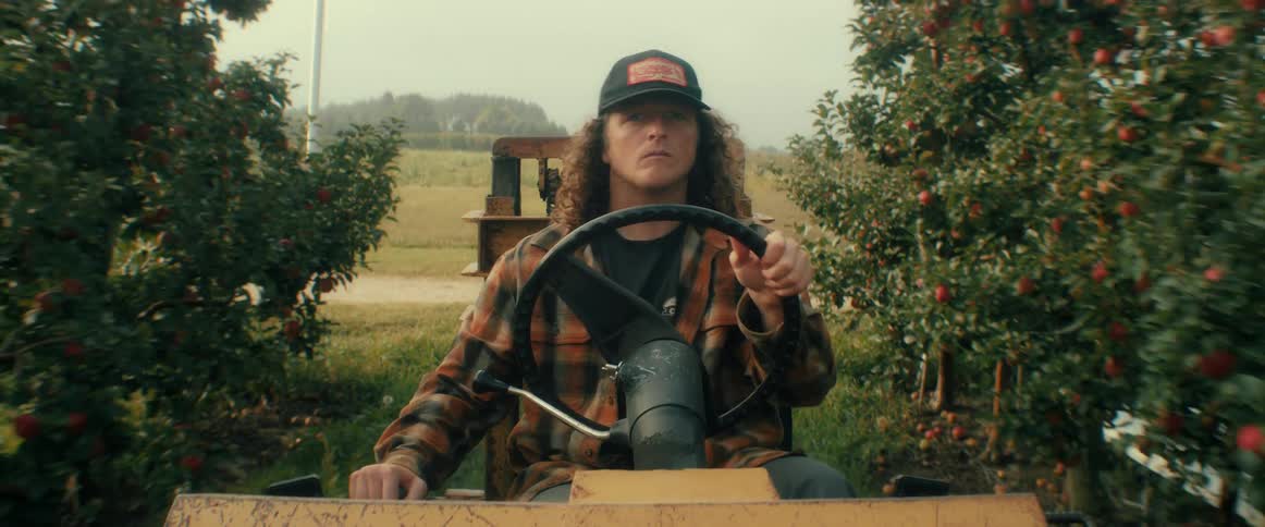 A young farmer in a flannel shirt and baseball cap drives a tractor through an apple orchard