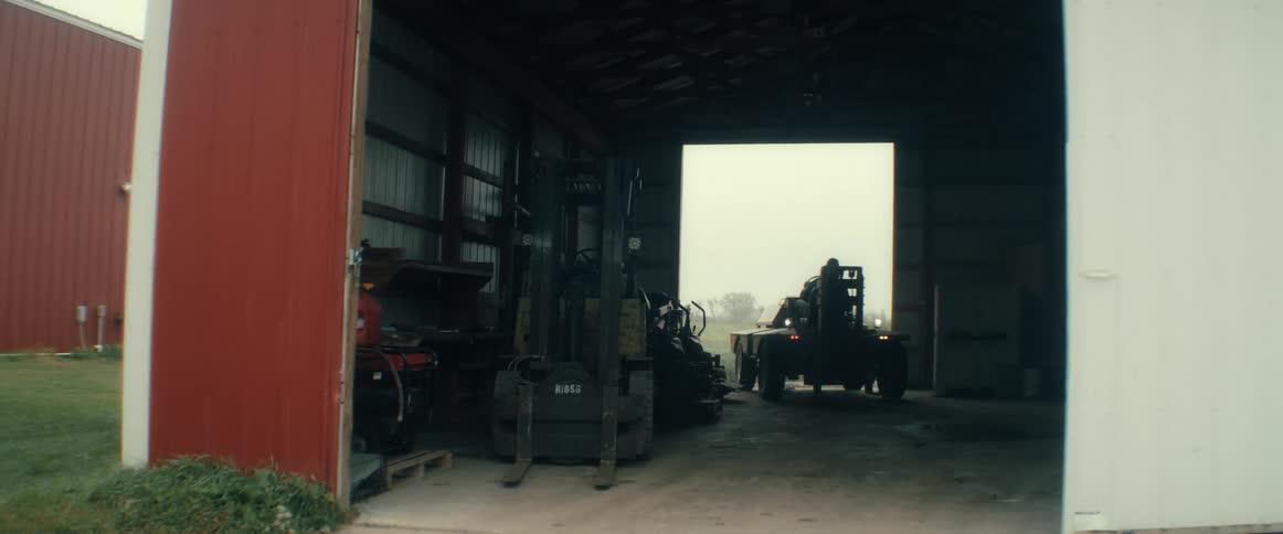 A forklift operator starts up and carefully backs a vehicle out of a dark rural barn onto a flatbed trailer on a misty day