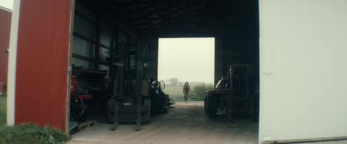 A man in a plaid jacket and hoodie walks from a foggy field into a dark industrial garage