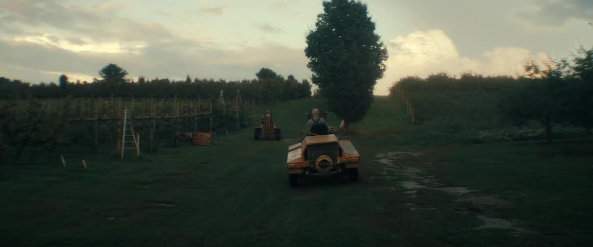 A farmer drives a unique yellow utility vehicle up a grassy hill through a rural orchard at dusk
