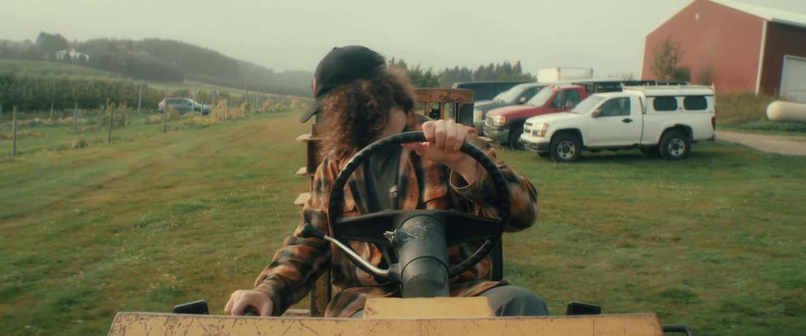 A smiling young man with long curly hair drives a tractor in a field on a rural farm