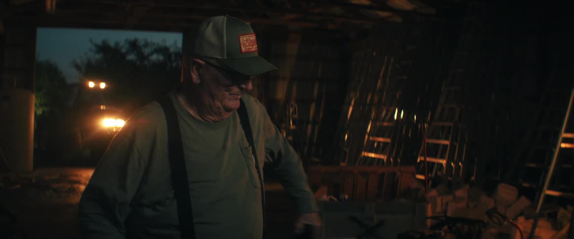 A portrait of a smiling elderly farmer in his barn at night, who then looks down to happily use his smartphone