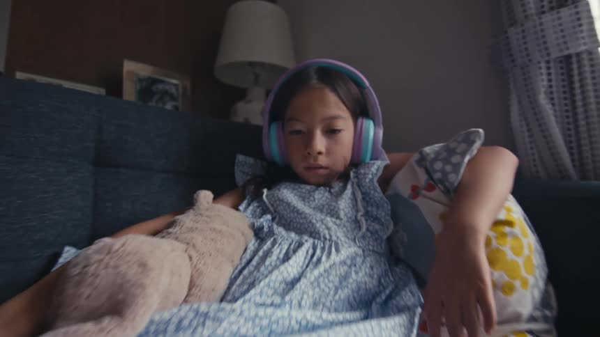 Young girl wearing colorful headphones relaxes on a couch while watching a tablet in a cozy living room